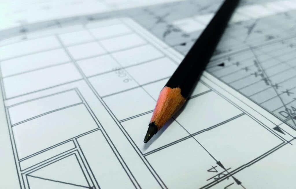 Ein angespitzter schwarzer Bleistift liegt auf einem Architekturplan mit feinen Linien und präzisen Maßen - ein Sinnbild für die sorgfältige Planung eines Fertighauses aus Holz, das nachhaltiges Bauen, Behaglichkeit und zeitloses Design vereint.