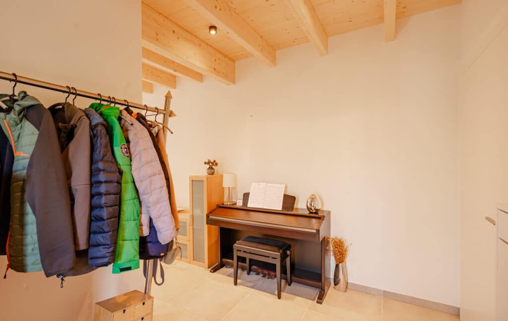 Ein heller, einladender Raum in einem nachhaltigen Holz-Fertighaus: Ein Klavier mit Notenständer und Hocker schafft musikalische Atmosphäre. An der Seite hängen Mäntel, auf dem Regal sorgen Trockenblumen und eine Lampe für wohnliche Akzente.