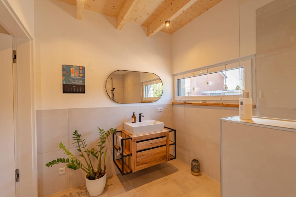 In diesem modernen Bad eines Hausbauprojekts bringt eine Decke aus Holz wohlige Wärme und natürliche Behaglichkeit. Großzügiger Spiegel, rechteckiges Waschbecken auf Holzwaschtisch, dezente Pflanzen und viel Tageslicht schaffen ein gemütliches Zuhausegefühl.