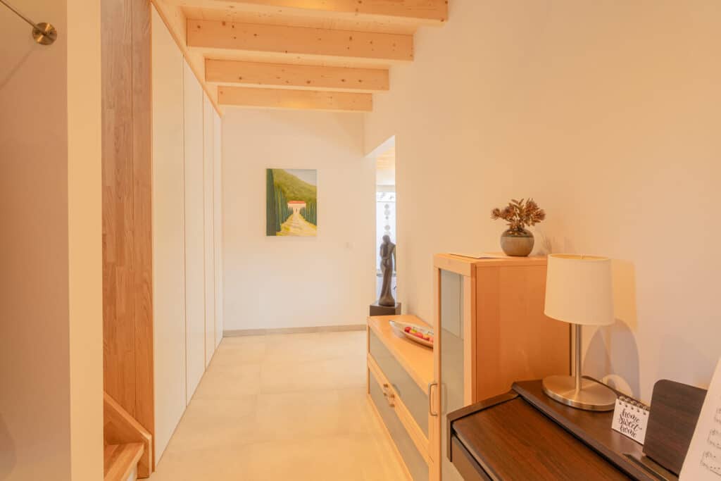 Eine freundliche Flur mit hellen Holzakzenten, sichtbaren Deckenbalken und stilvollen Details wie Lampe, Pflanze, Sideboard und Wandbild. Natürliches Licht durchflutet diesen behaglichen Raum - ein echtes Beispiel für modernes Holzhaus-Wohnen.