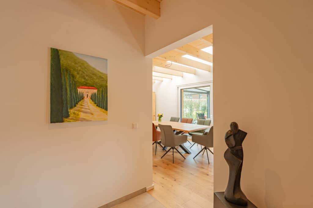 In einem lichtdurchfluteten Fertighaus aus Holz führt ein stilvoller Flur mit warmer Kunst und einer eleganten Skulptur zum offenen Essbereich. Natürliche Materialien, bunte Stühle und große Fenster verleihen dem Raum Behaglichkeit und Komfort.