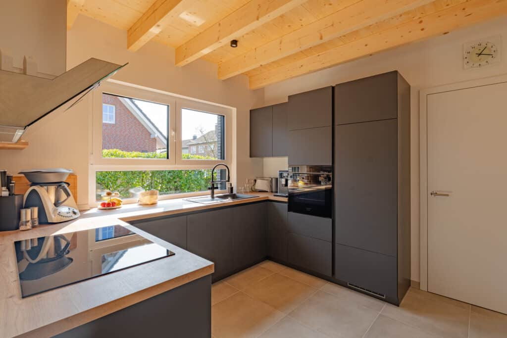 Gemütliche, moderne Holzküche im Fertighaus-Stil: Graue Schränke, integrierte Geräte und warme Holzarbeitsflächen treffen auf sichtbare Deckenbalken. Viel Tageslicht durch ein großes Fenster mit Blick ins Grüne schafft eine behagliche Atmosphäre.