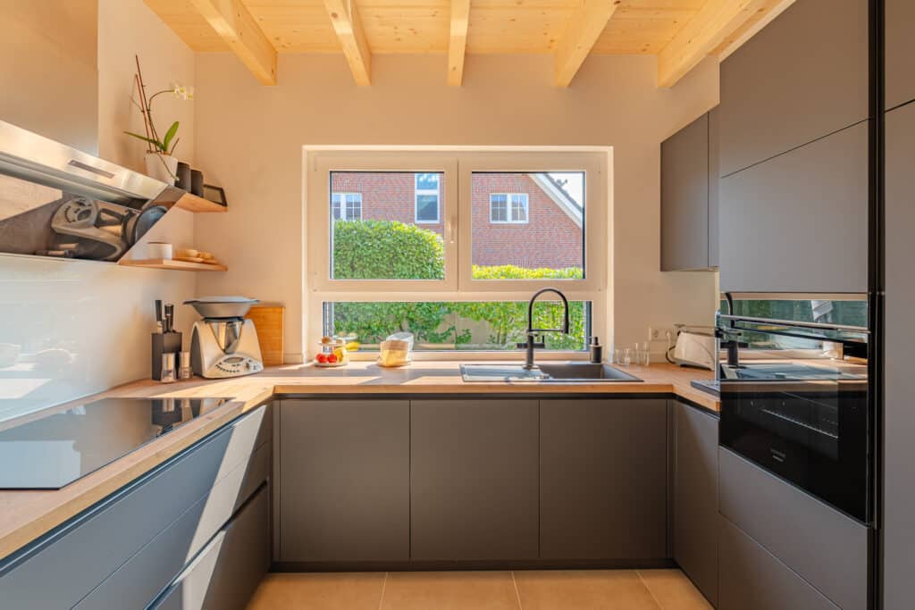 Moderne Küche mit grauen Schränken, warmen Holzarbeitsplatten und hochwertigen Einbaugeräten. Durch ein großes Fenster strömt Tageslicht herein und eröffnet den Blick ins Grüne - ein behaglicher Ort in Ihrem nachhaltigen Holz-Fertighaus.