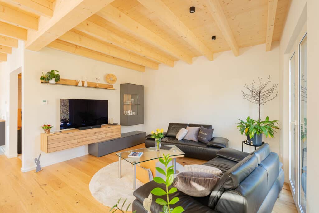 Ein gemütliches Wohnzimmer in einem Fertighaus aus Holz vereint moderne Eleganz mit nachhaltiger Bauweise: Schwarze Ledersofas, ein Glastisch und helle Holzböden sorgen für Wärme und Behaglichkeit, große Fenster und Pflanzen für lichtdurchflutete Wohnatmosphäre.