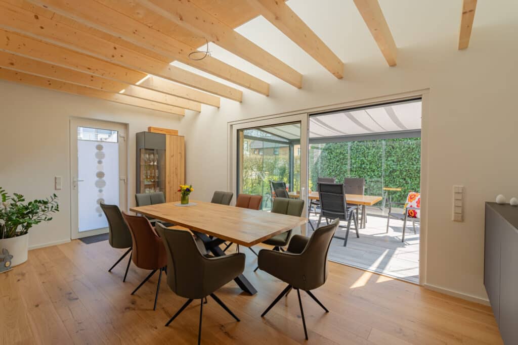 Helles Esszimmer im modernen Fertighaus aus Holz mit warmen Holzböden und sichtbaren Deckenbalken. Der Raum lädt mit einem Tisch für sechs Personen, großen Glas-Schiebetüren zur sonnigen Terrasse und Blick ins Grüne zum Wohlfühlen ein.