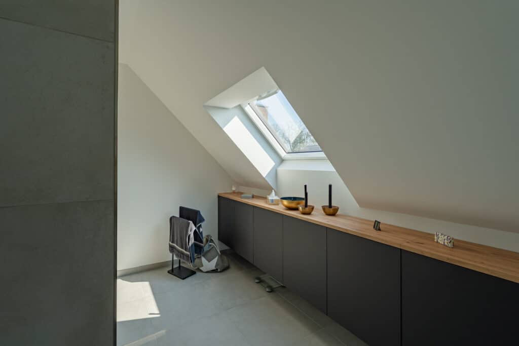 Ein modernes Dachgeschosszimmer mit schräger Decke und großzügigem Dachfenster. Die Kombination aus langem Holztresen und eleganten, schwarzen Unterschränken schafft ein behagliches Ambiente voller Licht und Wärme - perfekte Inspiration für Ihr Holzfertighaus.