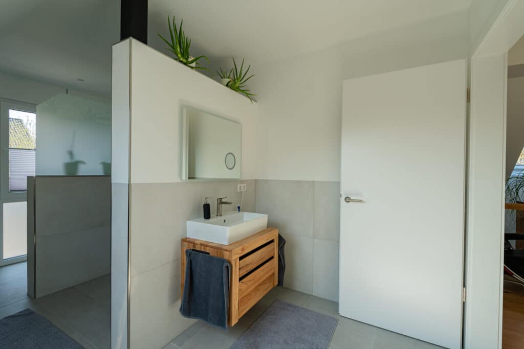 Modernes Bad mit schwebendem Waschtisch aus Holz, rechteckigem Becken, Spiegel und zwei kleinen Pflanzen auf einer Trennwand. Natürliche Materialien, sanftes Licht und behagliches Design schaffen Wohnkomfort im nachhaltigen Fertighaus aus Holz.