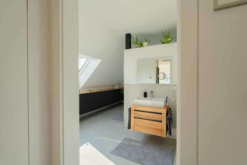 Helles, modernes Bad in einem Fertighaus aus Holz mit warmer Holz-Waschtischplatte, rundem Waschbecken und großem Spiegel. Helles Grau am Boden, dunkle Schränke, grüne Pflanzen auf dem Regal und natürliches Licht vom schrägen Dachfenster schaffen Wohnbehagen.