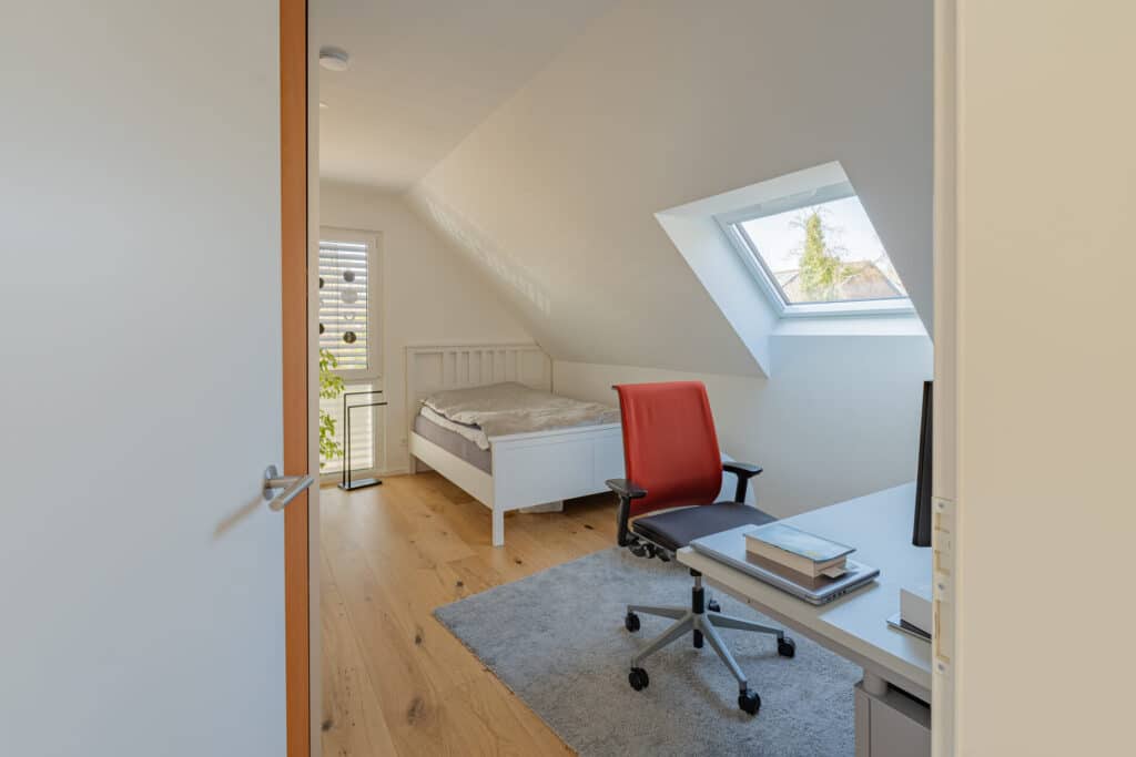 Ein helles, modernes Dachzimmer in einem nachhaltigen Fertighaus aus Holz vereint natürliche Materialien, eine gemütliche Atmosphäre und zeitgemäßes Design: schräges Dach, Einzelbett, weißer Schreibtisch mit Bildschirm, roter Stuhl und großes Fenster für viel Licht.