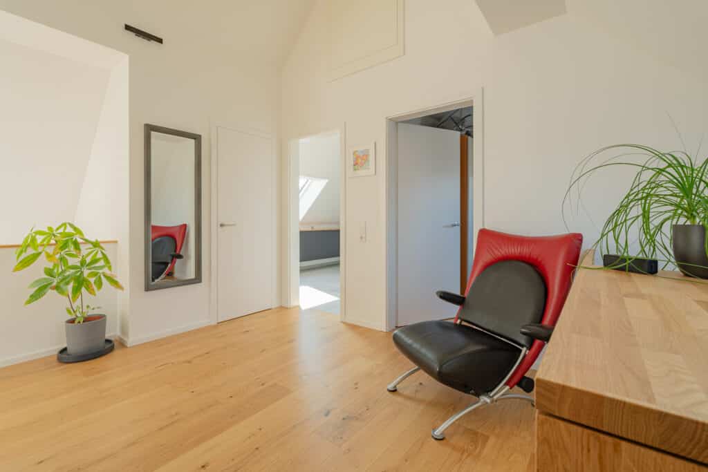 Heller, moderner Raum in einem Fertighaus aus Holz mit hellem Holzboden und weißen Wänden. Ein großer Spiegel, eine Zimmerpflanze, ein rot-schwarzer Stuhl und ein Schreibtisch schaffen eine behagliche Atmosphäre. Die offene Tür führt in einen sonnigen Nebenraum.