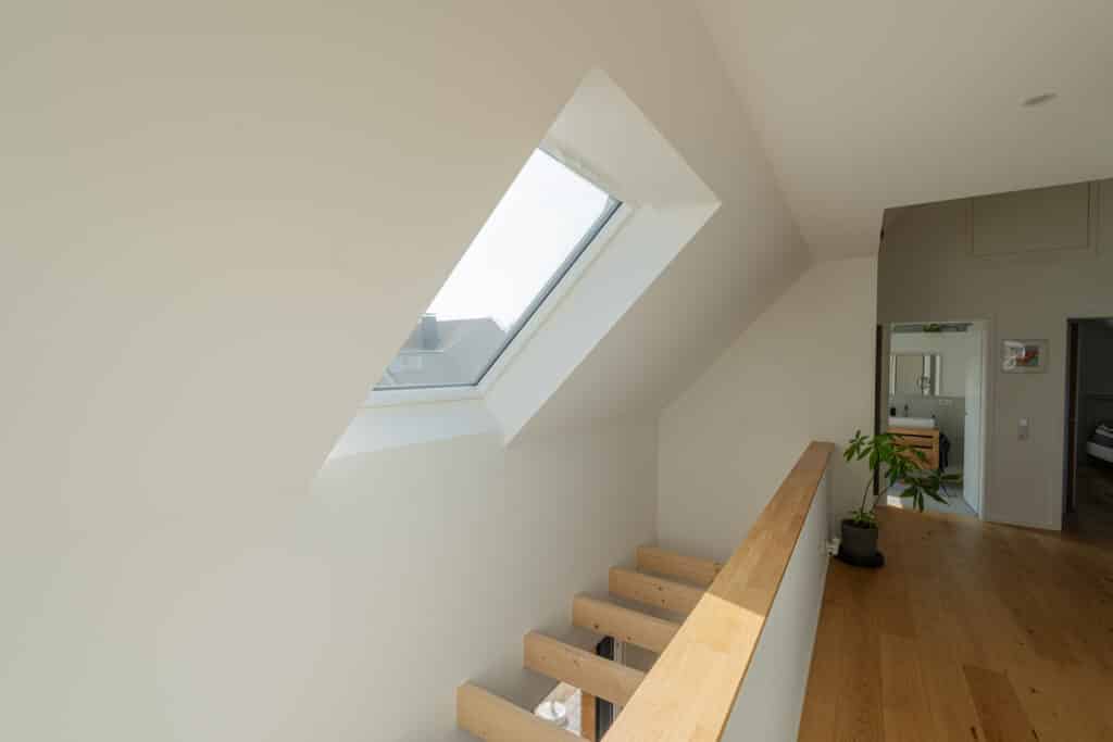 Eine lichtdurchflutete Flur mit warmen Holzböden zeigt modernes Fertighaus-Design: Schräges Dachfenster, stilvolles Geländer und Blick ins gemütliche Zimmer mit Zimmerpflanze. Natürliches Licht und nachhaltige Materialien schaffen Wohlfühlatmosphäre.