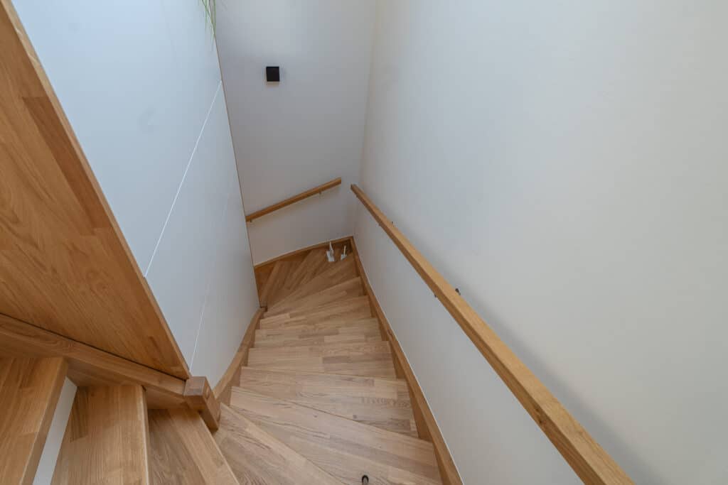 In einem hellen, behaglichen Fertighaus aus Holz führen stilvoll geschwungene Treppen mit Handläufen beidseits durch eine weiße, einladende Flur. Am Fuß der Treppe sitzt eine kleine weiße Katze und verleiht dem Zuhause eine warme, wohnliche Atmosphäre.