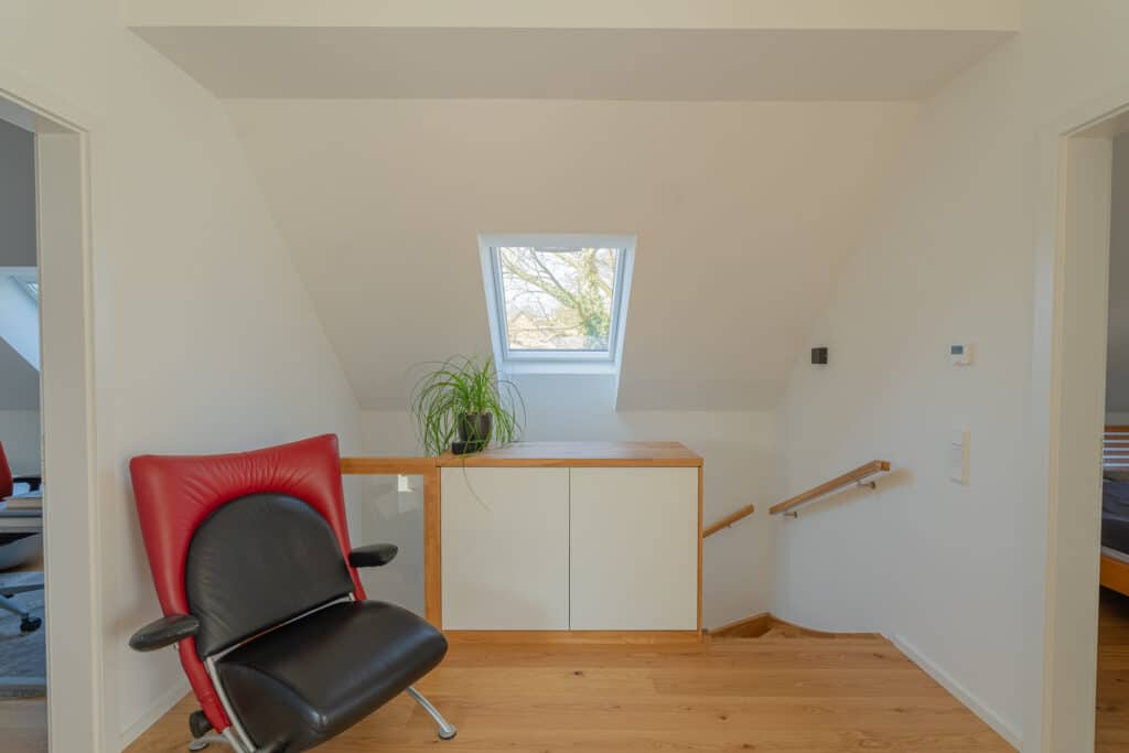 Ein lichtdurchfluteter, moderner Raum mit warmen Holzböden, dezenten Wänden sowie einer stilvollen Kombination aus schwarzem und rotem Stuhl, kleinem Schrank und dekorativer Zimmerpflanze - ein inspirierendes Beispiel für nachhaltiges Wohlfühlwohnen.