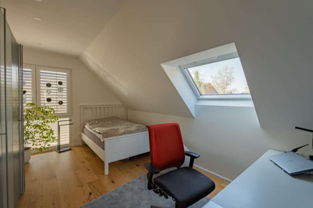 Ein lichtdurchflutetes, modernes Dachgeschoss-Schlafzimmer in einem nachhaltigen Holz-Fertighaus lädt mit Behaglichkeit und stilvollem Design zum Wohlfühlen ein: Einzelbett, Schreibtisch mit Laptop und gemütlichem Stuhl, großer Schrank, Pflanze und großzügigem Fenster.