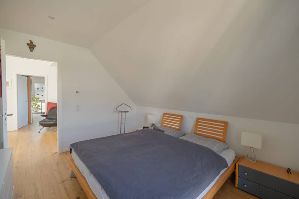 Ein lichtdurchflutetes Schlafzimmer mit schrägen weißen Decken in einem modernen Fertighaus aus Holz strahlt Behaglichkeit und Wärme aus. Natürliche Materialien, ein Holz-Bett mit grauer Bettwäsche und viel Sonnenlicht schaffen Wohlfühlatmosphäre.