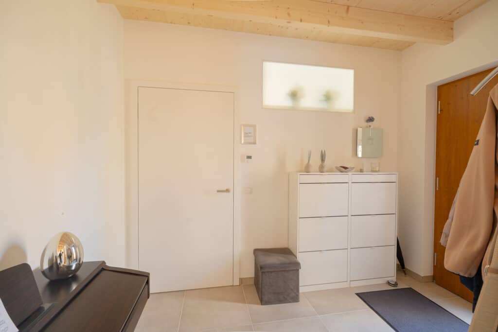 Lichterfüllter, minimalistischer Eingangsbereich mit weißer Tür, weißem Schuhschrank, beigen Wänden und Holzdecke. Natürliche Materialien und zeitloses Design schaffen ein warmes Ambiente - perfekte Inspiration für nachhaltiges Wohnen im Fertighaus aus Holz.