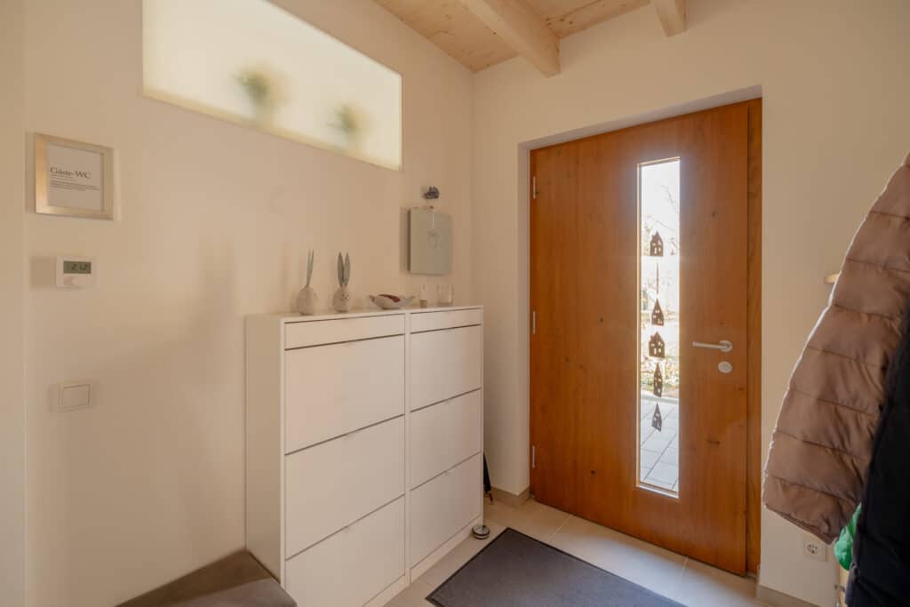 Ein heller Eingangsbereich mit einer stilvollen Holztür samt Glaseinsätzen und lichtdurchflutetem Fenster lädt zum Ankommen ein. Die weiße Schuhkommode, liebevolle Dekofiguren und natürliche Materialien unterstreichen die Behaglichkeit. Ein Mantel hängt rechts.