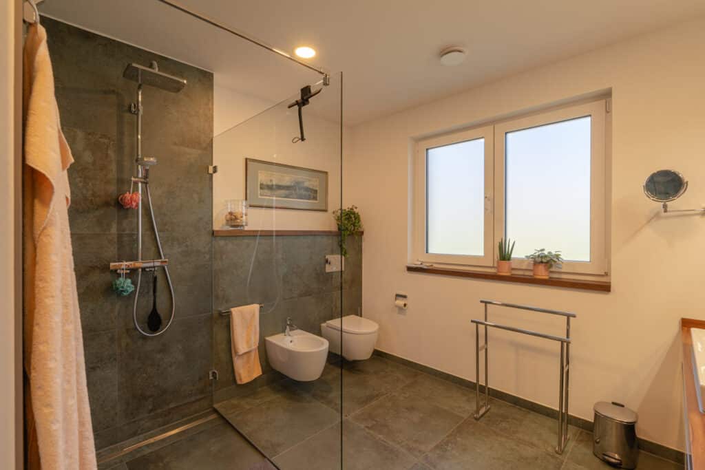 Dieses moderne Bad mit bodengleicher Glasdusche, dunklen Fliesen, Bidet und großem Fenster überzeugt durch stilvolle Behaglichkeit. Natürliche Materialien und liebevolle Details wie Pflanzen schaffen eine Wohlfühloase in Ihrem Fertighaus aus Holz.