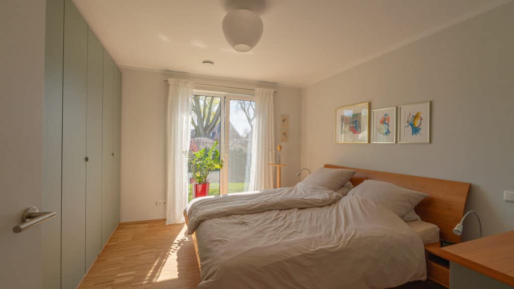 Ein lichtdurchflutetes Schlafzimmer in einem modernen Fertighaus aus Holz verbindet natürliche, helle Holzböden mit hochwertiger Einrichtung: Ein gemütliches Doppelbett mit beiger Bettwäsche, ein geräumiger Schrank und stimmungsvolle Kunst über dem Bett schaffen eine behagliche Atmosphäre. Sonnenlicht fällt durch transparente Vorhänge auf einen roten Blumentopf am Fenster - ein Ort zum Wohlfühlen.