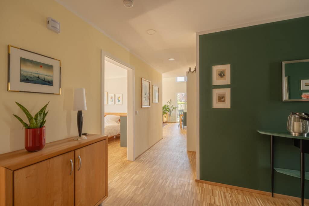 Ein heller Flur mit Holzfußboden, sanft gelb-grünen Wänden und liebevoll arrangierten Details wie gerahmter Kunst und einer Holzkredenz mit Zimmerpflanze strahlt wohnbehagliche Wärme aus. Geöffnete Türen lassen Licht ins stilvoll gemütliche Zuhause fluten.