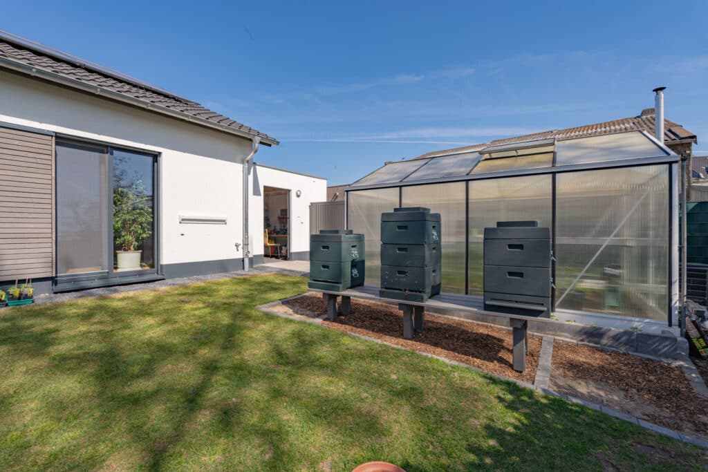 Ein einladender Garten mit üppigem Rasen, eleganten Wegen und einem modernen Fertighaus aus Holz mit großen Fenstern. Ein Gewächshaus sowie liebevoll arrangierte Bienenstöcke unterstreichen die nachhaltige Wohlfühlatmosphäre.