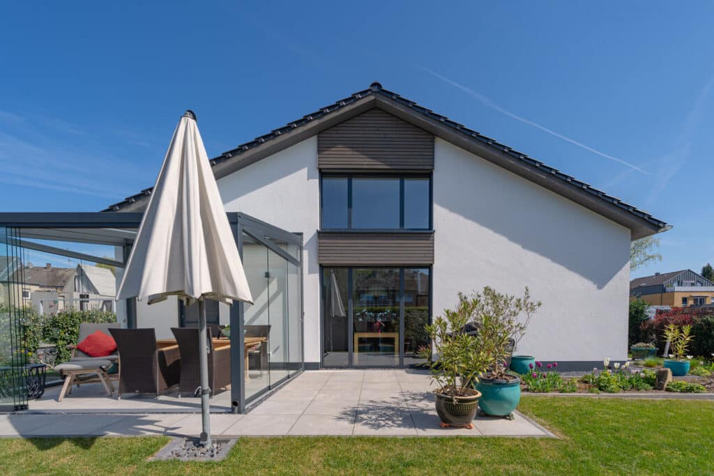 Modernes Holz-Fertighaus mit geneigtem Dach und großen Fenstern, umgeben von einer einladenden Terrasse mit gemütlichen Outdoor-Möbeln, liebevoll platzierten Pflanzen und einem geschlossenen weißen Sonnenschirm - unter strahlend blauem Himmel.