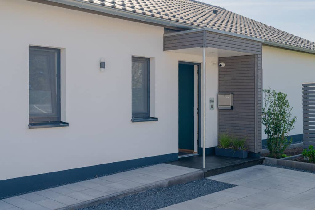 Einladender Eingang eines modernen Holz-Fertighauses mit blauer Tür, zwei Fenstern und lichtgrauen Wänden. Kleine Pflanzen und ein gepflegter, gefliester Weg verleihen Wärme und Wohnlichkeit - Qualität, die Geborgenheit für Generationen schafft.