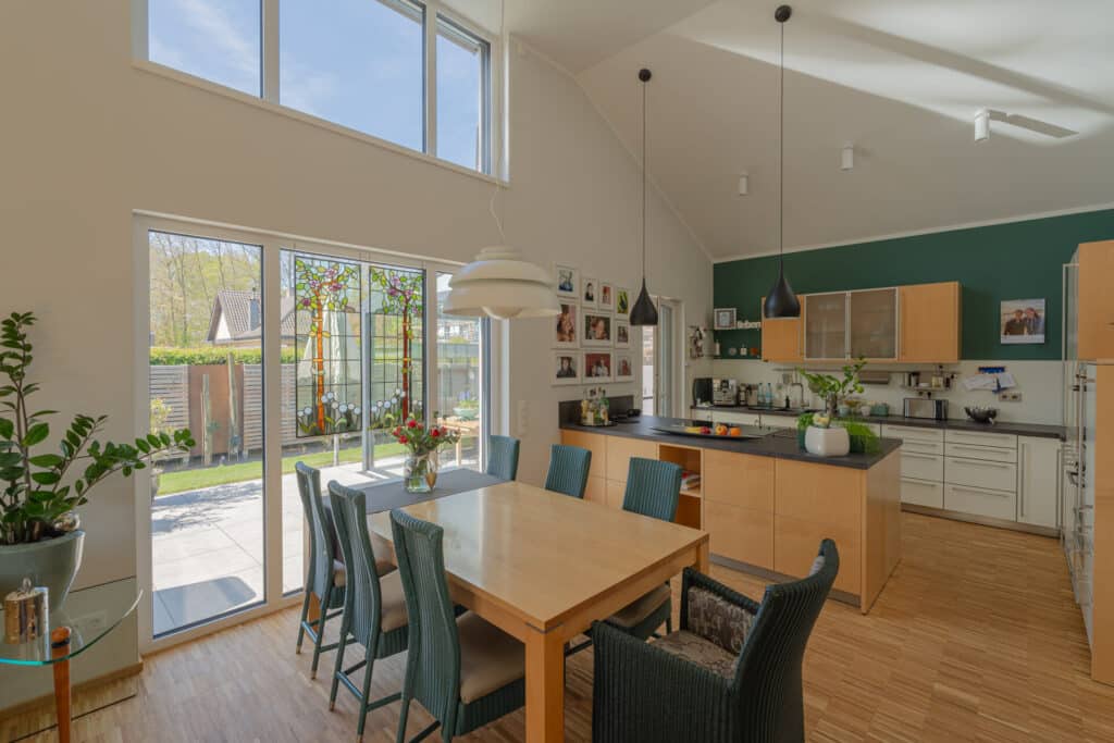 Helles, modernes Ess- und Küchenareal mit großen Fenstern, Holztisch und Stühlen, grüner Akzentwand, stilvollen Hängelampen und üppigen Pflanzen. Der Blick in den Garten vermittelt Behaglichkeit - perfekte Inspiration für nachhaltiges Holzhaus-Bauen.