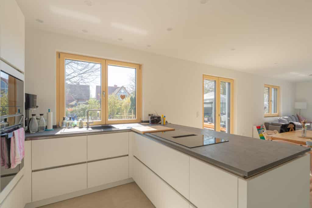 Im Mittelpunkt dieses nachhaltigen Fertighauses aus Holz lädt eine helle, moderne Küche mit weißen Schränken und grauer Arbeitsplatte zum Wohlfühlen ein. Große Fenster schenken viel Tageslicht und bieten Blick ins Grüne, Wohn- und Essbereich schließen sich an.