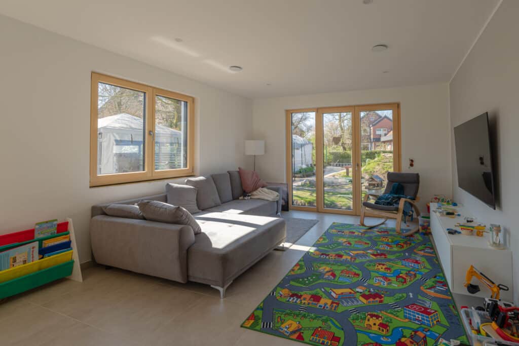 Ein lichtdurchflutetes Wohnzimmer in einem nachhaltigen Fertighaus aus Holz schafft mit einer grauen Wohnlandschaft, praktischem Spielzeug-Organizer, gemütlichem Sessel und stilvoller Beleuchtung eine behagliche Atmosphäre mit Blick in den Garten.