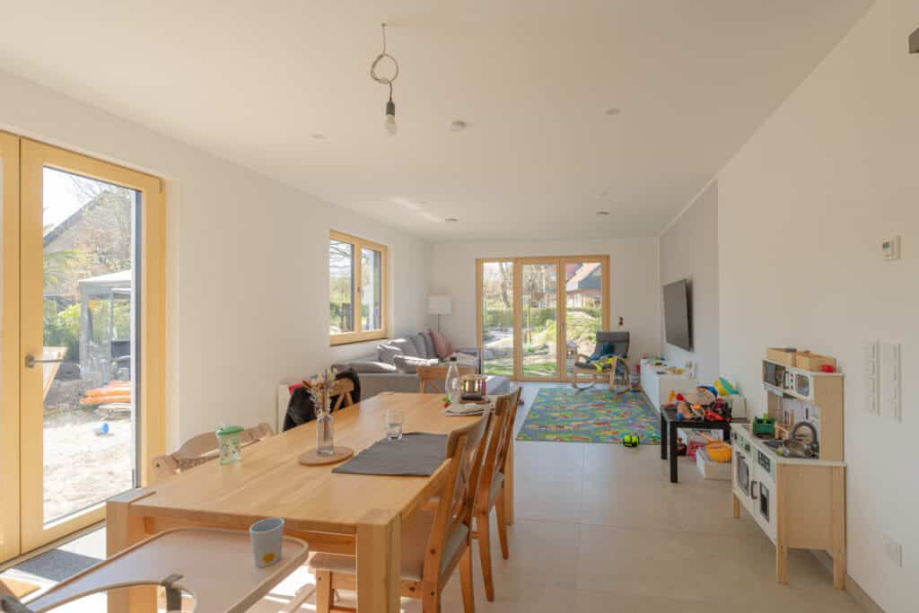 Modernes Wohn- und Esszimmer in einem nachhaltigen Fertighaus aus Holz, ausgestattet mit natürlichen Holzmöbeln, Kinderspielzeug und Spielküche. Große Fenster schaffen helles Ambiente und geben den Blick ins Grüne frei. Eine gemütliche Sofa-Ecke lädt zum Entspannen ein.