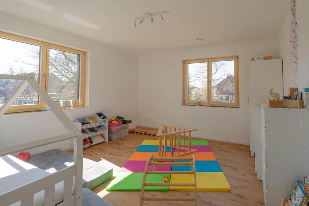 Heller, einladender Spielraum in einem modernen Fertighaus aus Holz: Holzboden, bunte Schaumstoffmatten und eine niedrige Kletterstruktur schaffen Behaglichkeit. Spielzeugregale und große Fenster sorgen für Licht und wohnliches Ambiente.
