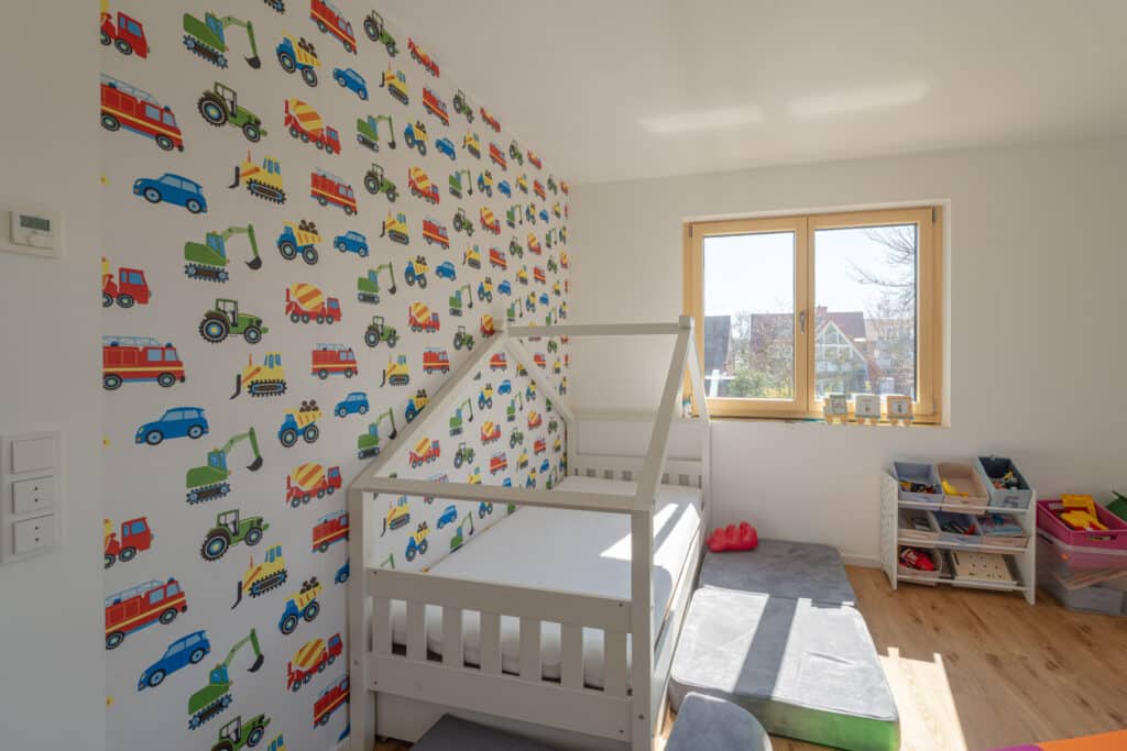 Ein liebevoll gestaltetes, sonnendurchflutetes Kinderzimmer mit einem weißen Holzhaus-Bett, fröhlicher Fahrzeugtapete und Spielzeugregalen - ein behaglicher Rückzugsort voller Inspiration für kleine Bauherren und Träumer nachhaltiger Fertighäuser.