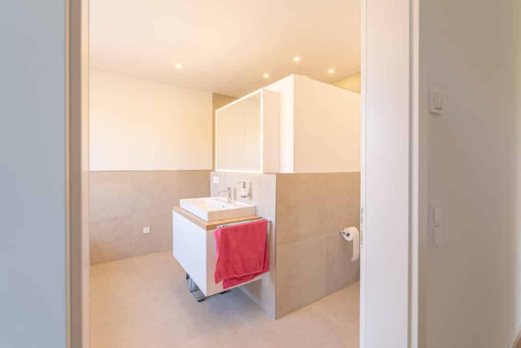 Dieses moderne Bad in einem Fertighaus aus Holz strahlt mit seinen beigen Fliesen an Wand und Boden, einem weißen Waschbecken samt Spiegel, rotem Handtuch und stilvollen Details behagliche Wärme und hochwertige, nachhaltige Bauweise aus.