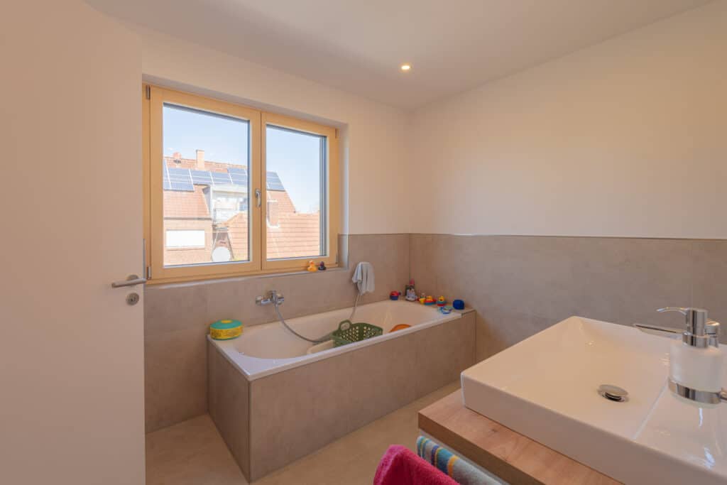 Ein modernes Badezimmer im nachhaltigen Holz-Fertighaus überzeugt mit behaglicher Atmosphäre: Rechteckige Badewanne unter dem Fenster, Kinder-Badespielzeug auf dem Wannenrand, Waschbecken mit Seifenspender auf Holz, beige Fliesen.
