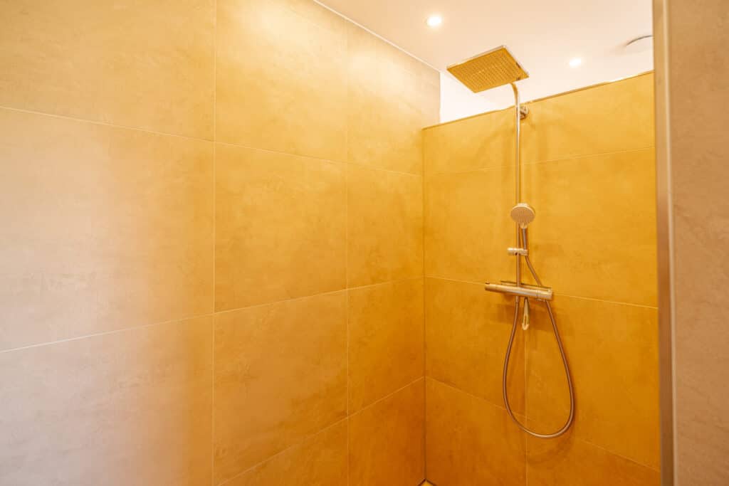 Helle, behagliche Walk-in-Dusche mit großen beigen Fliesen, wandmontiertem Regenduschkopf und Handbrause. Das minimalistische Design wird durch natürliche Materialien und energiesparende Deckenleuchten unterstrichen - ideal fürs Holz-Fertighaus.