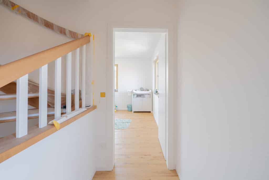 Heller Flur mit Holztreppe links, teils von Schutzmaterial umhüllt, führt in einen sonnendurchfluteten Raum mit hellen Holzböden - einladend und modern. Durch eine offene Tür ist ein Wickeltisch sichtbar - ideale Wohlfühlatmosphäre fürs Zuhause.