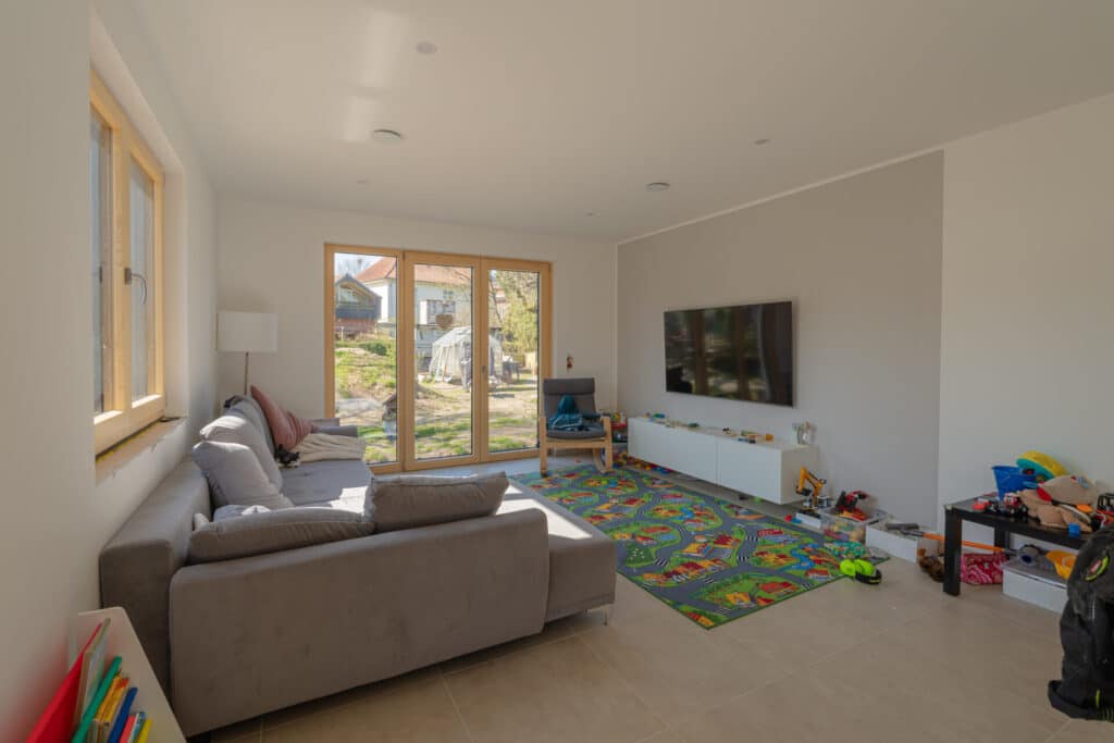 Ein helles Wohnzimmer in einem modernen Fertighaus aus Holz lädt mit einer grauen Wohnlandschaft, großen Fenstern und viel Tageslicht zum Verweilen ein. Der warme Holzboden, die freundliche Spielecke und hochwertige Materialien sorgen für behagliche Wohlfühlatmosphäre.