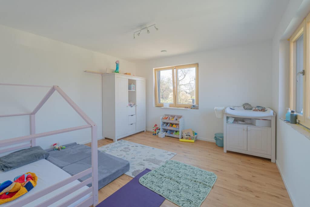 Dieses helle, aufgeräumte Kinderzimmer mit Holzfußboden, weißen Wänden und großen Fenstern verströmt wohnliche Wärme und Komfort. Natürliche Materialien schaffen einen behaglichen Raum mit Bodenbett, Wickeltisch, Schrank und Spielmatten.