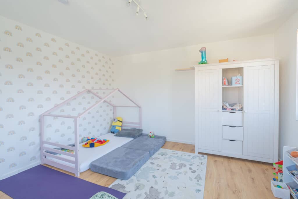 Ein helles, aufgeräumtes Kinderzimmer mit Hausbett aus Holz vermittelt Wärme und Geborgenheit - ideal für Ihr nachhaltiges Fertighaus. Natürliche Materialien, weiße Schränke, bunte Spielsachen und Regenbogentapete sorgen für wohnliches Wohlfühlambiente.