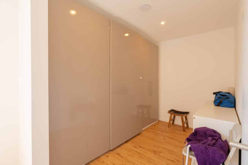 Ein moderner, minimalistischer Ankleideraum in einem nachhaltigen Fertighaus aus Holz: Beige Hochglanzschränke, warmer Holzfußboden, eine weiße Bank mit blauem Tasche und lila Handtuch sowie ein Hocker mit schwarzem Sitz sorgen für Wohlfühlatmosphäre.