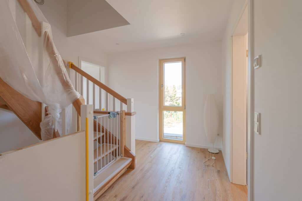 Lichtdurchfluteter Flur eines nachhaltigen Holzhaus-Projekts mit hellen Holzböden, einer durch Folie geschützten Treppe und einem weißen Kindergitter. Natürliches Licht sowie hochwertige Materialien schaffen eine einladende, behagliche Atmosphäre.