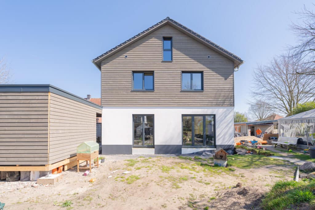 Ein modernes, zweistöckiges Holzhaus mit grauer Fassade und großzügigen Fenstern verspricht lichtdurchflutete Räume und behagliches Wohnklima. Links steht ein kleiner Holzschuppen, im Garten lockert eine natürliche Rasenfläche unter blauem Himmel die Szenerie auf.