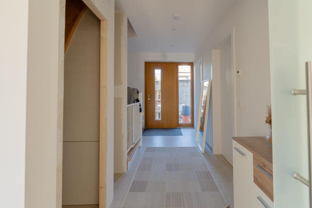 Ein heller Flur mit weißen Wänden, gemustertem Boden und einer Holz-Eingangstür mit Glaseinsätzen vermittelt zeitlose Behaglichkeit. Natürliche Materialien und moderne Details wie Spiegel und Vase sorgen für ein wohnliches, nachhaltiges Ambiente.