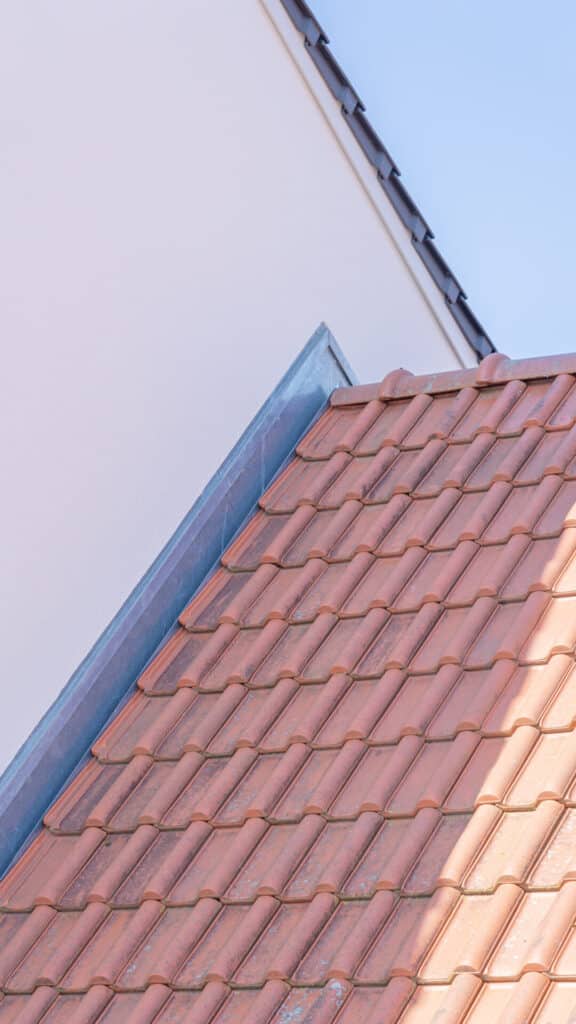 Die Nahaufnahme eines geneigten Daches mit rotbraunen Ziegeln an einer weißen Hauswand vor strahlend blauem Himmel zeigt, wie moderner Holzbau mit natürlichen Materialien zu einem behaglichen und zeitlosen Zuhause voller Wärme inspiriert.