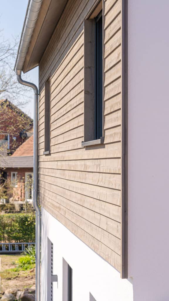 Ein modernes Fertighaus aus Holz im Seitenausschnitt: Die obere Etage zeigt warme Holzverkleidung, darunter weiße Putzfassade. Ein großes Fenster und eine Metall-Dachrinne betonen das nachhaltige, behagliche Design. Im Hintergrund steht ein weiteres Haus.