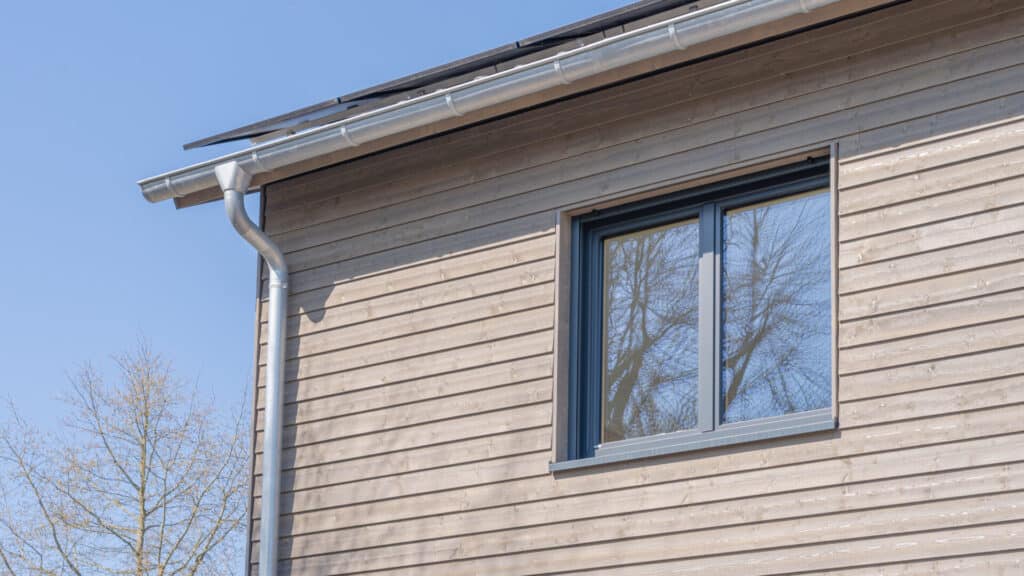 Detailaufnahme der Außenfassade eines modernen Fertighauses aus Holz mit horizontaler Holzverkleidung, großem Fenster im grauen Rahmen, Regenrinne und Solarpaneelen auf dem Dach - ein Beispiel für nachhaltige Bauweise mit Wohlfühlatmosphäre unter blauem Himmel.