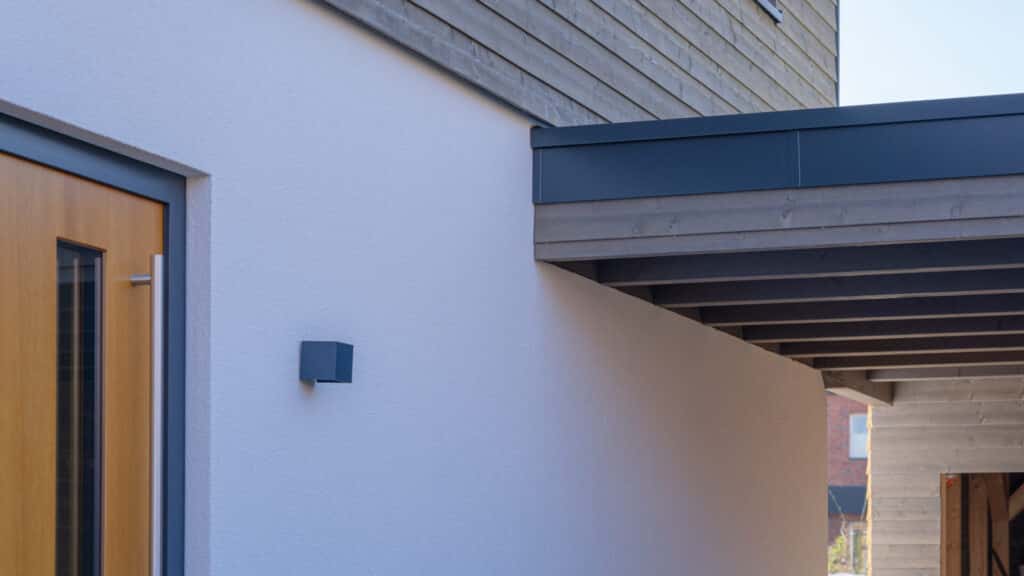 Einladendes modernes Haus mit weißer Putzfassade, Holztür samt Glaseinsatz, grauem Vordach und offenen Balken - kombiniert nachhaltige Bauweise und zeitgemäßes Design. Dieses Fertighaus aus Holz strahlt Wärme, Komfort und Behaglichkeit aus.