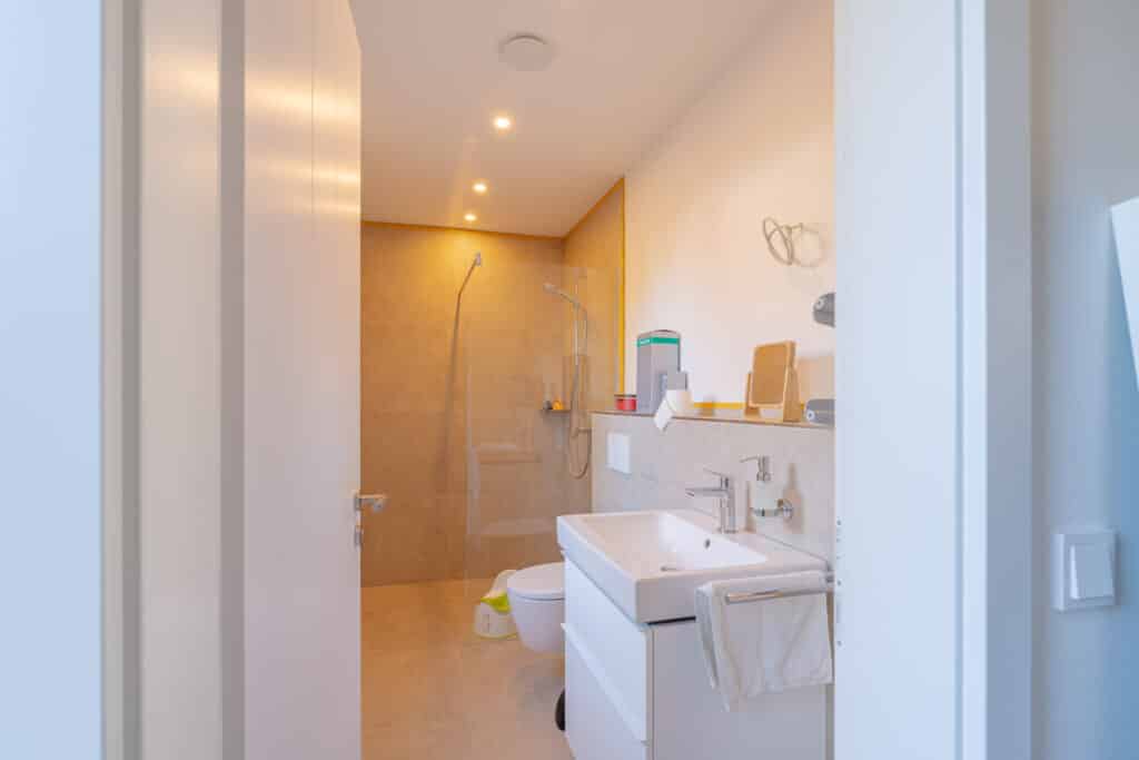 Ein einladendes, modernes Bad in einem Holz-Fertighaus mit sandfarbenen Fliesen, bodengleicher Dusche, weißer Toilette und stilvoller Waschtischanlage. Offene Regale schaffen Platz für Pflegeprodukte; Handtuch und grüner Mülleimer setzen wohnliche Akzente.