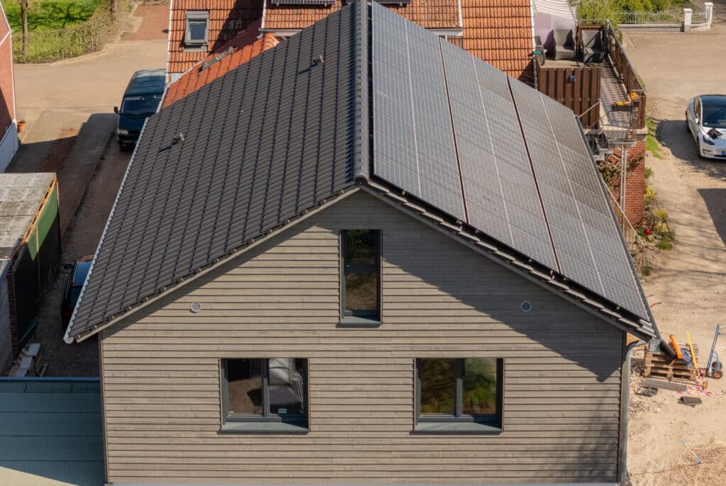 Ein modernes Fertighaus aus Holz mit grauem Ziegeldach und großzügig integrierten Solarpanels präsentiert sich in nachhaltiger Bauweise. Die wohnliche Holzfassade, helle Fenster sowie umgebende Häuser und ein geparktes Auto unterstreichen das behagliche Wohngefühl.